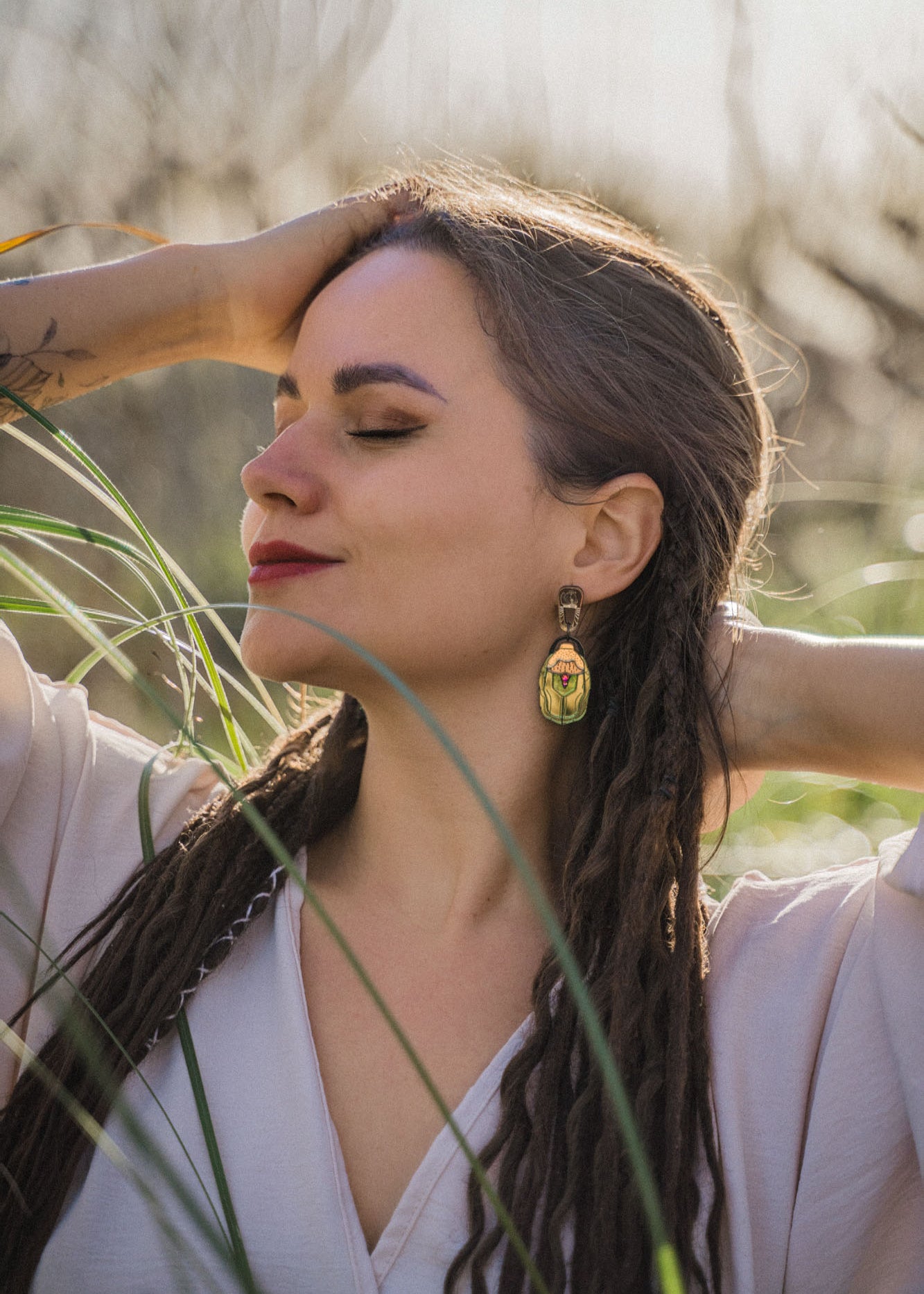 A woman with long, braided hair and bold Rose Chafer Earrings stands outdoors with her eyes closed, smiling softly and raising her arms behind her head in a relaxed, nature-inspired setting among tall grass.
