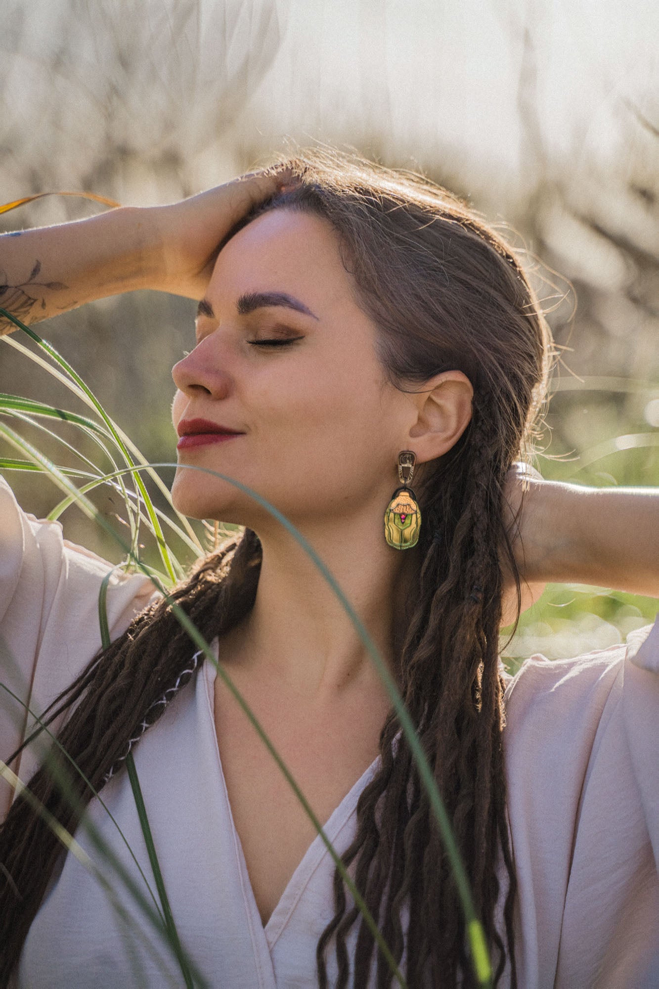 A woman with long, braided hair and bold Rose Chafer Earrings stands outdoors with her eyes closed, smiling softly and raising her arms behind her head in a relaxed, nature-inspired setting among tall grass.