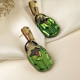 A pair of handcrafted gold and green Rose Chafer Earrings with an abstract face design, featuring purple and black accents, displayed on a cream-colored fabric background with dried flowers in the corner.