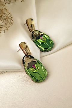 A pair of handcrafted gold and green Rose Chafer Earrings with an abstract face design, featuring purple and black accents, displayed on a cream-colored fabric background with dried flowers in the corner.