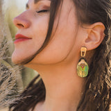 A woman with closed eyes and red lipstick faces the sunlight, wearing a large, nature-inspired earring shaped like a matryoshka doll. Her long brown hair is styled in loose dreadlocks.