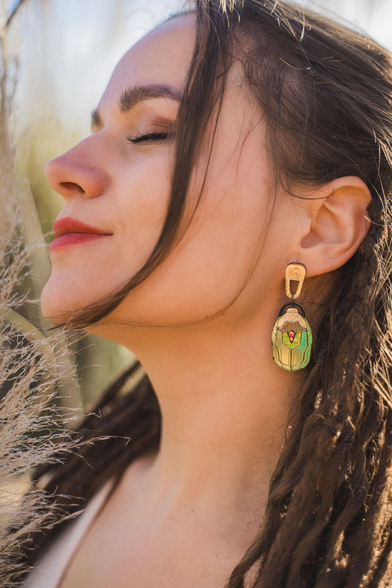 A woman with closed eyes and red lipstick faces the sunlight, wearing a large, nature-inspired earring shaped like a matryoshka doll. Her long brown hair is styled in loose dreadlocks.