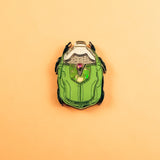A green and black enamel Rose Chafer Pin, resembling a sci-fi or robot helmet, is centered on a plain peach background—a meaningful design perfect for lovers of handcrafted accessories.