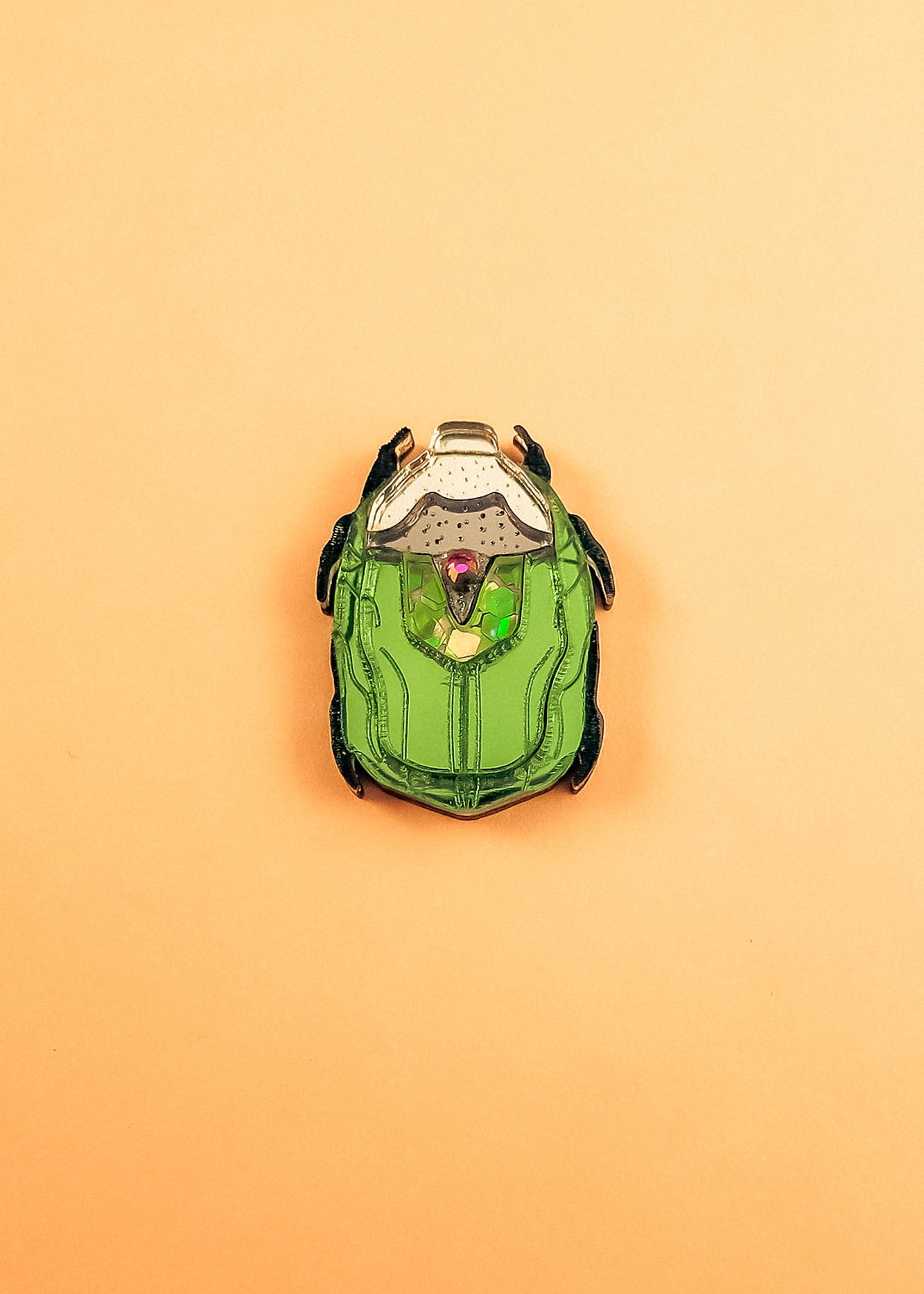 A green and black enamel Rose Chafer Pin, resembling a sci-fi or robot helmet, is centered on a plain peach background—a meaningful design perfect for lovers of handcrafted accessories.