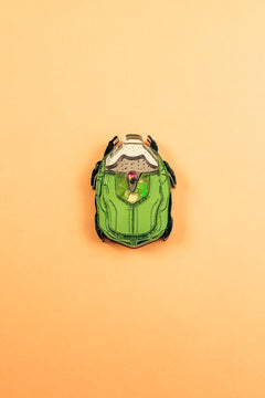 A green and black enamel Rose Chafer Pin, resembling a sci-fi or robot helmet, is centered on a plain peach background—a meaningful design perfect for lovers of handcrafted accessories.