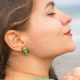 A woman with closed eyes and a slight smile faces sideways, wearing a green top and nature-inspired earrings, with the beach and ocean blurred in the background.