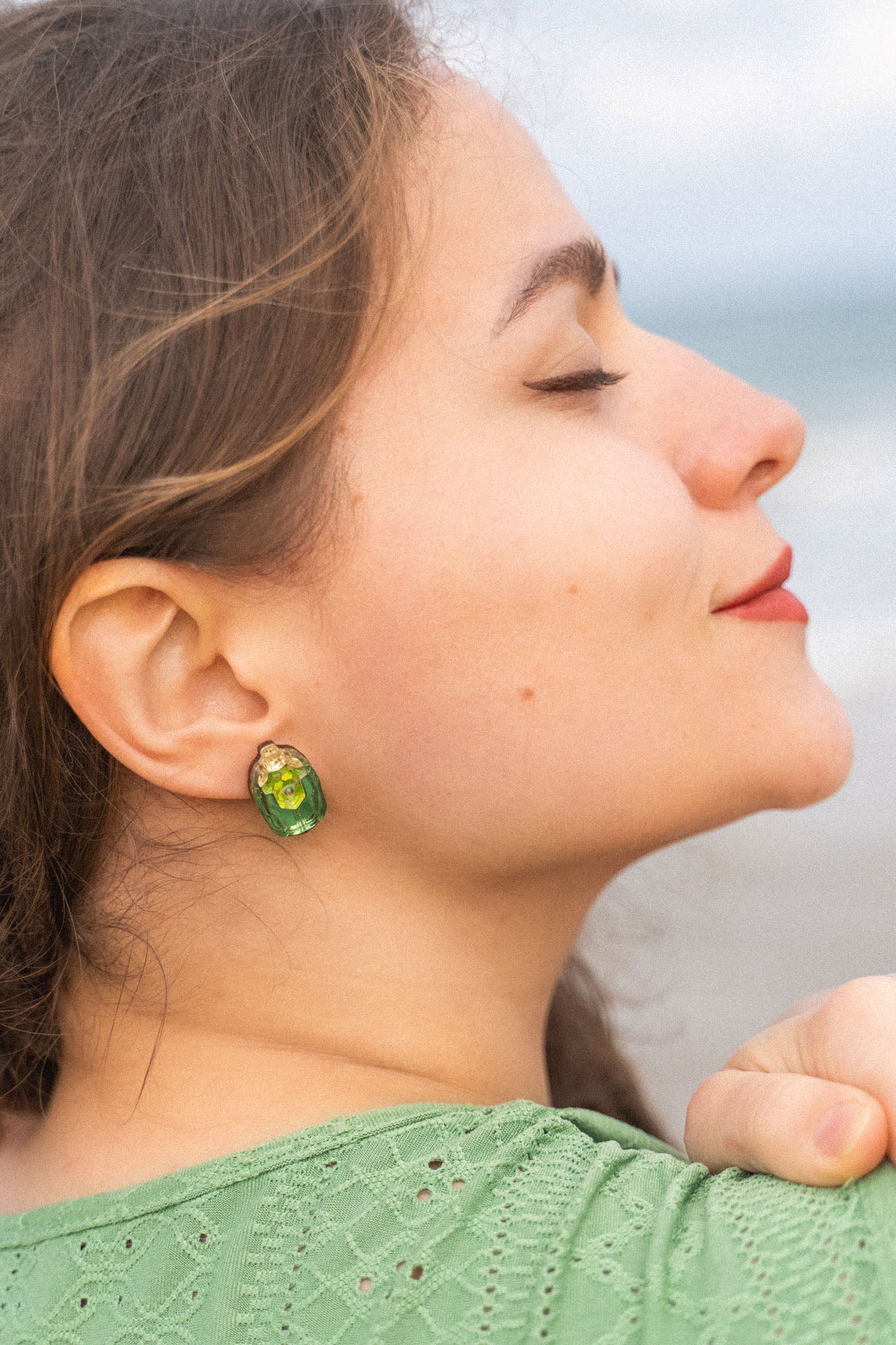 A woman with closed eyes and a slight smile faces sideways, wearing a green top and nature-inspired earrings, with the beach and ocean blurred in the background.