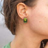 Close-up of a person wearing Rose Chafer Studs—nature-inspired earrings with green gemstones and gold details. The person has wavy brown hair, a green top, and is set against a softly blurred background.