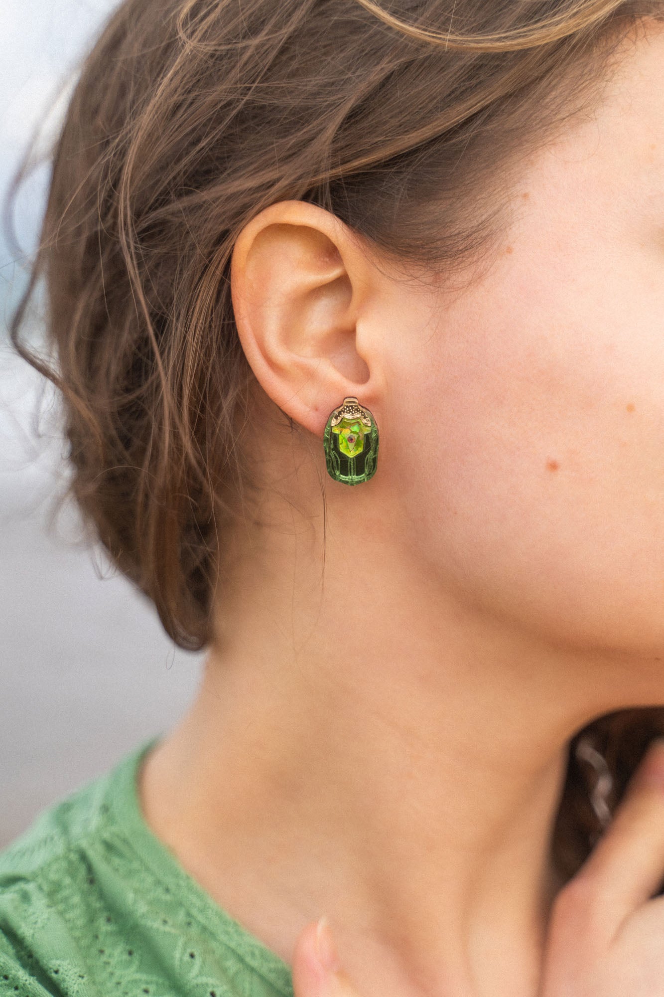 Close-up of a person wearing Rose Chafer Studs—nature-inspired earrings with green gemstones and gold details. The person has wavy brown hair, a green top, and is set against a softly blurred background.