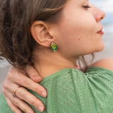 A woman with closed eyes and a peaceful expression rests her hand on her shoulder. She wears a green, eyelet-patterned top and handcrafted earrings. Her brown hair is slightly tousled as she stands outdoors.