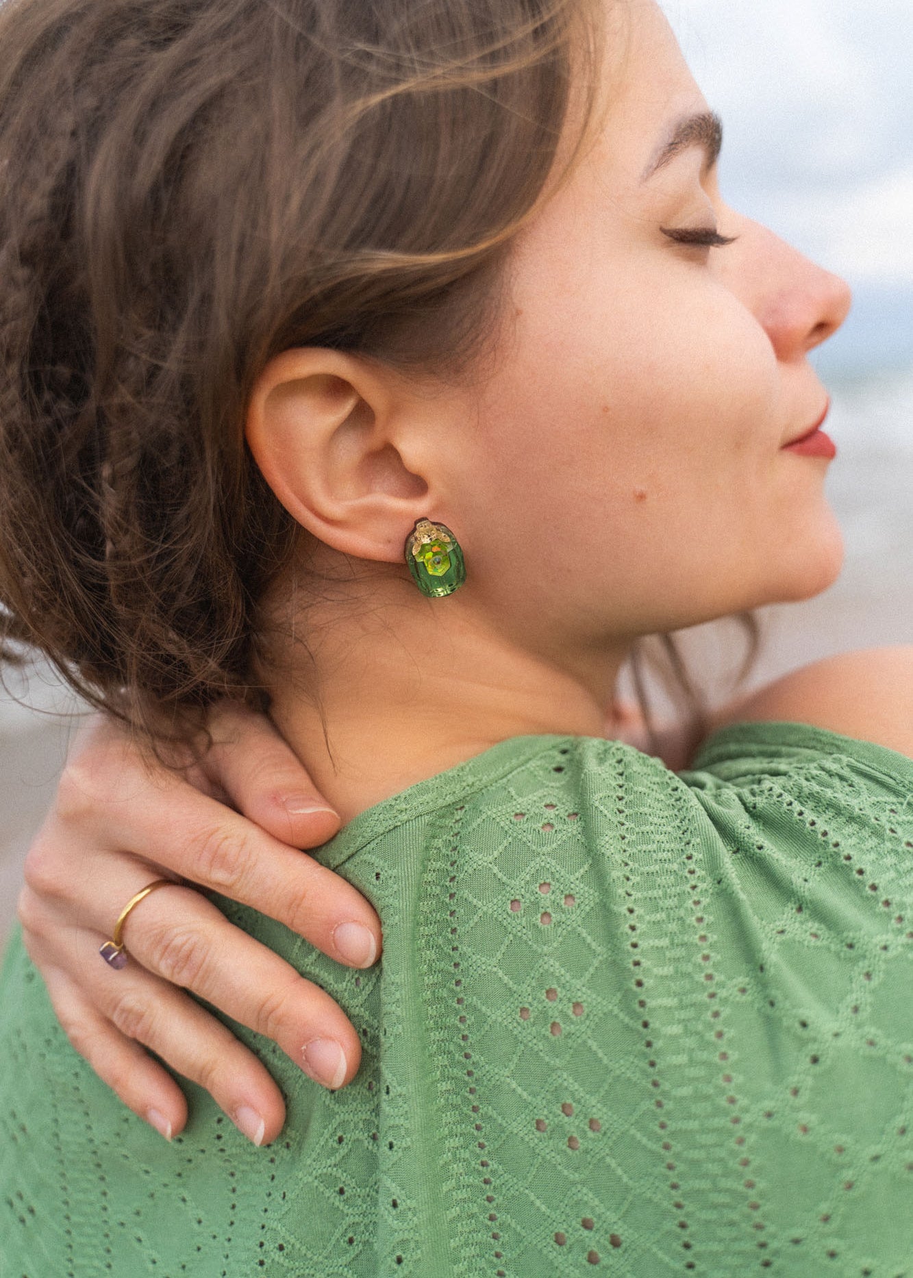 A woman with closed eyes and a peaceful expression rests her hand on her shoulder. She wears a green, eyelet-patterned top and handcrafted earrings. Her brown hair is slightly tousled as she stands outdoors.