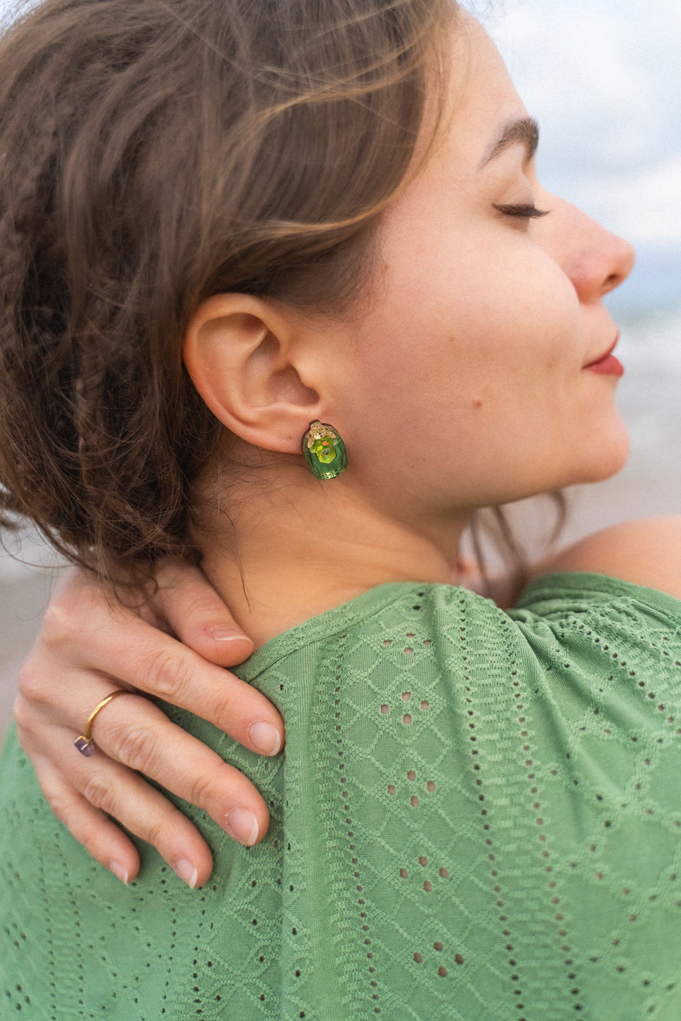 A woman with closed eyes and a peaceful expression rests her hand on her shoulder. She wears a green, eyelet-patterned top and handcrafted earrings. Her brown hair is slightly tousled as she stands outdoors.