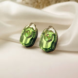 A pair of green, faceted gemstone Rose Chafer Studs with metallic tops displayed on white satin fabric, these nature-inspired earrings shine against a soft, blurred background.