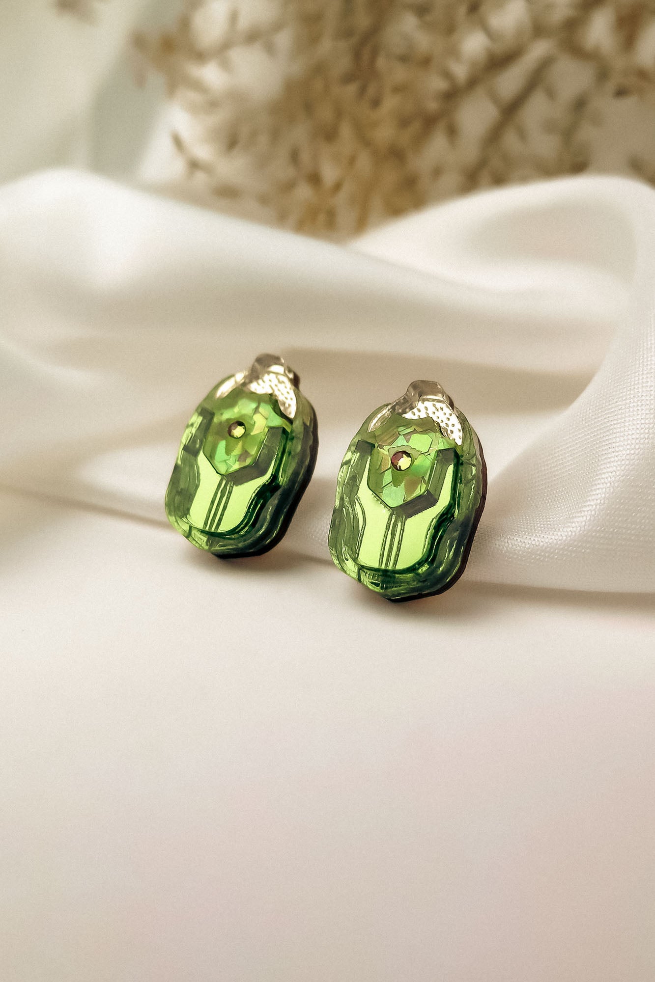 A pair of green, faceted gemstone Rose Chafer Studs with metallic tops displayed on white satin fabric, these nature-inspired earrings shine against a soft, blurred background.