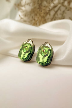A pair of green, faceted gemstone Rose Chafer Studs with metallic tops displayed on white satin fabric, these nature-inspired earrings shine against a soft, blurred background.