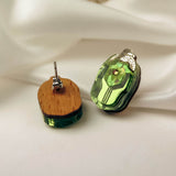 A pair of handcrafted Rose Chafer Studs with green translucent stones and wooden backs are displayed on white satin fabric. One nature-inspired earring shows the front, while the other reveals the wooden back and metal post.