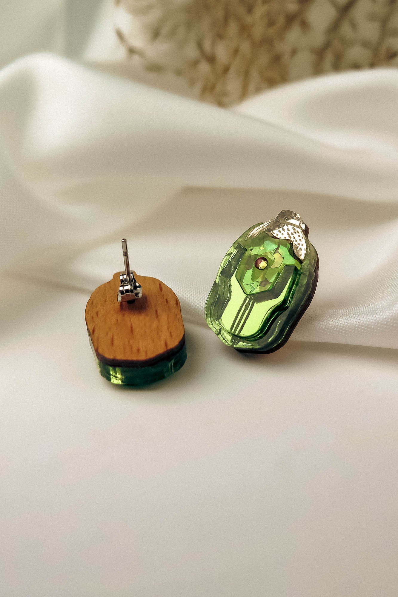 A pair of handcrafted Rose Chafer Studs with green translucent stones and wooden backs are displayed on white satin fabric. One nature-inspired earring shows the front, while the other reveals the wooden back and metal post.