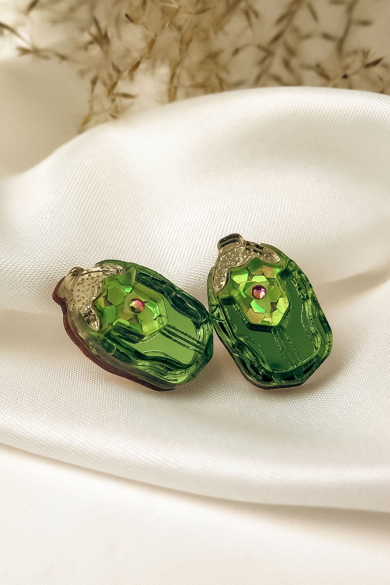 Two green, oval-shaped glass craft beads with a faceted surface and a small pink center gem, perfect for nature-inspired earrings, resting on a white silky fabric backdrop with blurred dried flowers in the background.