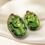 A close-up of Rose Chafer Studs, handcrafted earrings featuring green gemstones with gold accents, resting on soft white satin. Their shiny, faceted surface and decorative top make them perfect for nature lovers.
