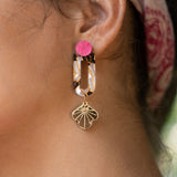 Close-up of a womans ear wearing Sakura Earrings—a statement earring with a bright pink stud, tortoiseshell oval link, and gold floral charm. A pink patterned headscarf adds an extra touch of elegance and artistry to the look.