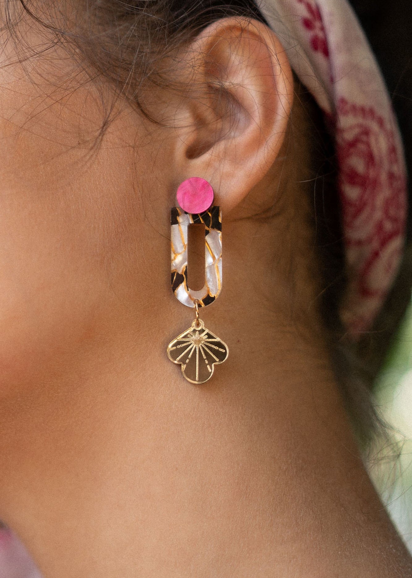 Close-up of a womans ear wearing Sakura Earrings—a statement earring with a bright pink stud, tortoiseshell oval link, and gold floral charm. A pink patterned headscarf adds an extra touch of elegance and artistry to the look.