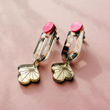 The Sakura Earrings showcase elegance and artistry with pink circular studs, gold rectangular links, and dangling flower-shaped charms, all displayed on a soft pink surface.
