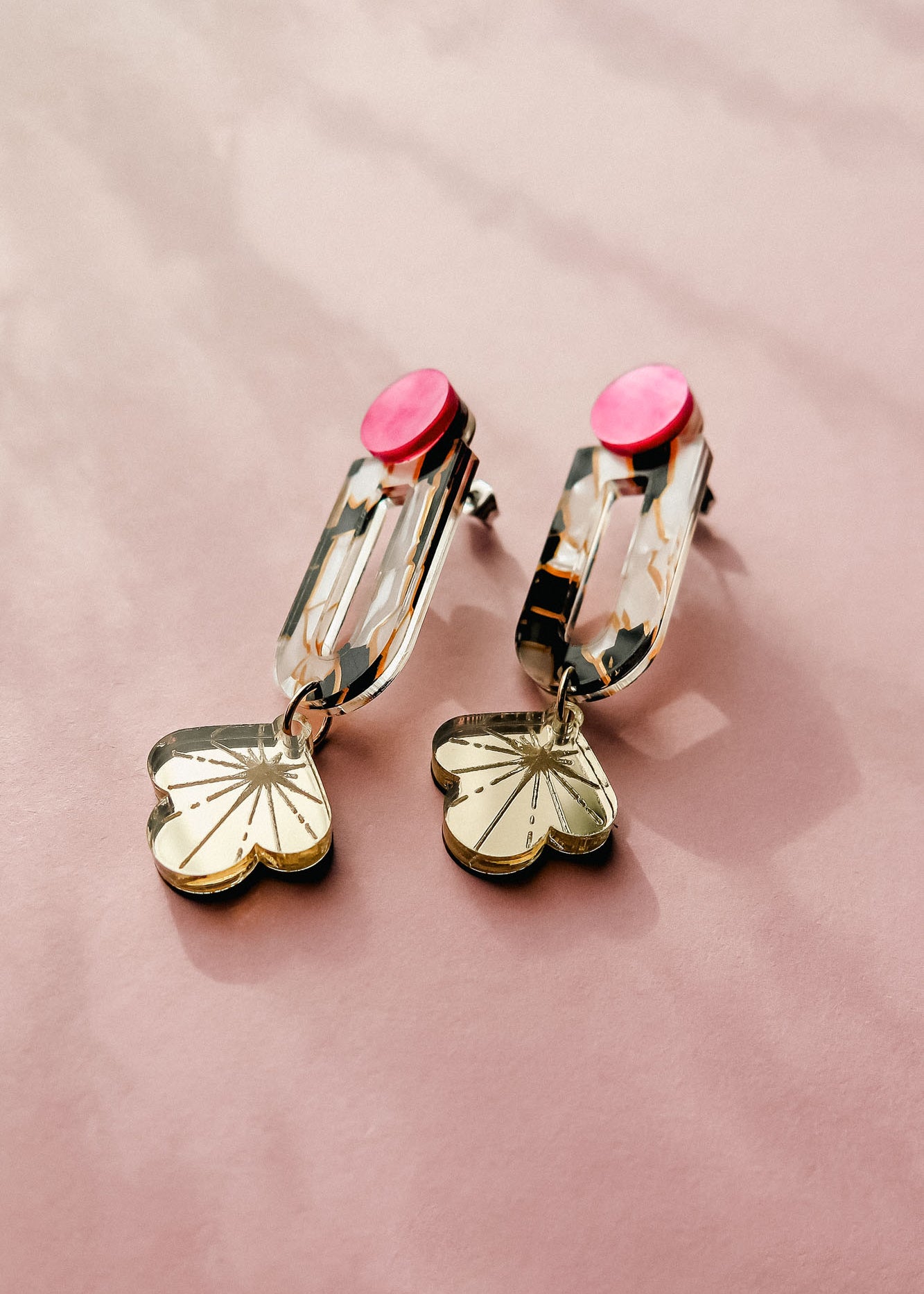 The Sakura Earrings showcase elegance and artistry with pink circular studs, gold rectangular links, and dangling flower-shaped charms, all displayed on a soft pink surface.