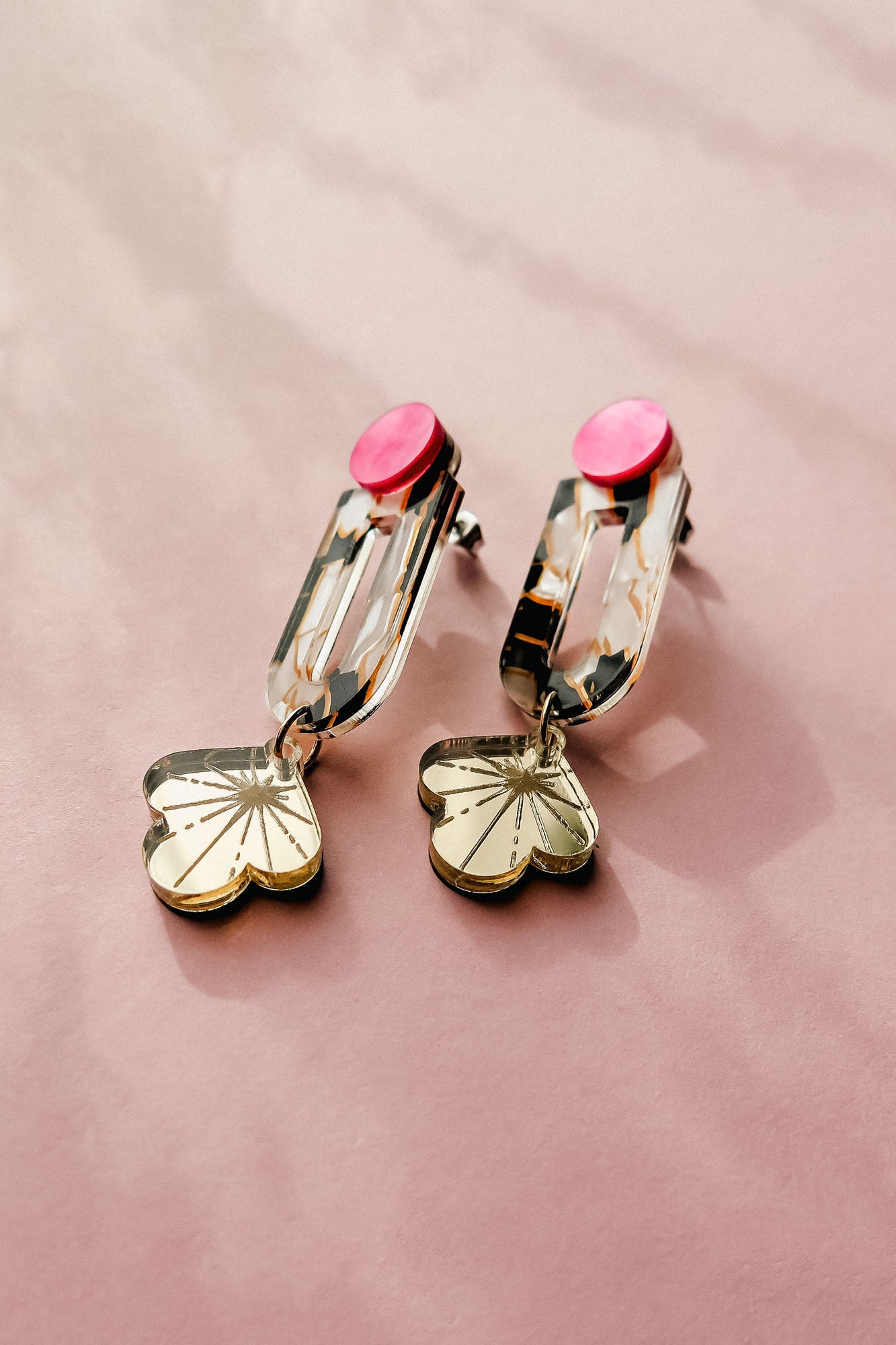 The Sakura Earrings showcase elegance and artistry with pink circular studs, gold rectangular links, and dangling flower-shaped charms, all displayed on a soft pink surface.