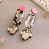 Showcasing elegance and artistry, the Sakura Earrings feature pink circular studs, marbled white and gold rectangular links, and gold flower-shaped pendants, all displayed on light beige fabric with dried plant accents.