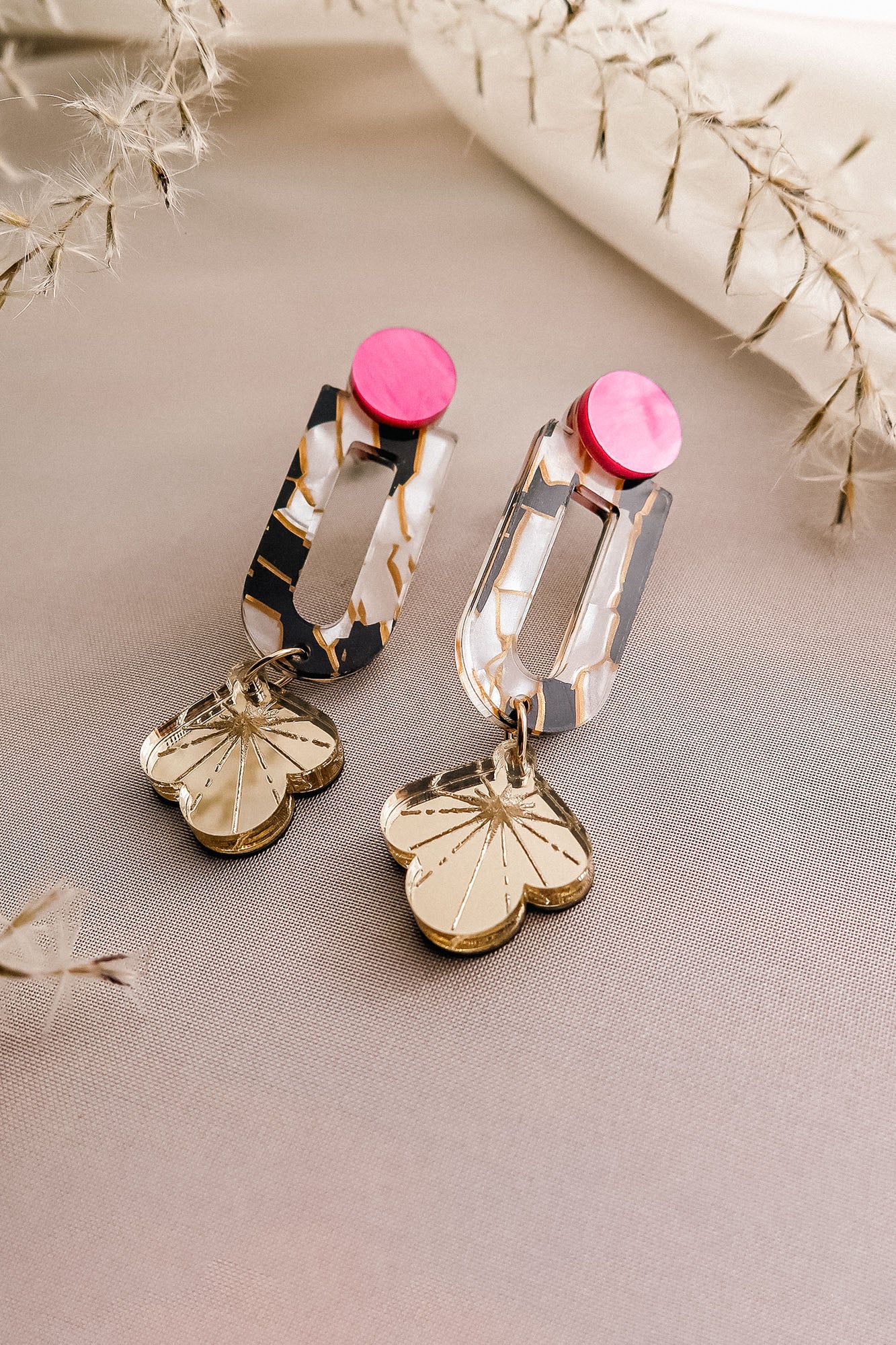 Showcasing elegance and artistry, the Sakura Earrings feature pink circular studs, marbled white and gold rectangular links, and gold flower-shaped pendants, all displayed on light beige fabric with dried plant accents.