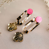 A pair of modern Sakura Earrings featuring pink circular studs, rectangular tortoiseshell links, and dangling gold heart-shaped charms with etched starburst designs, displayed on a light beige fabric background—an expression of artistry and elegance.