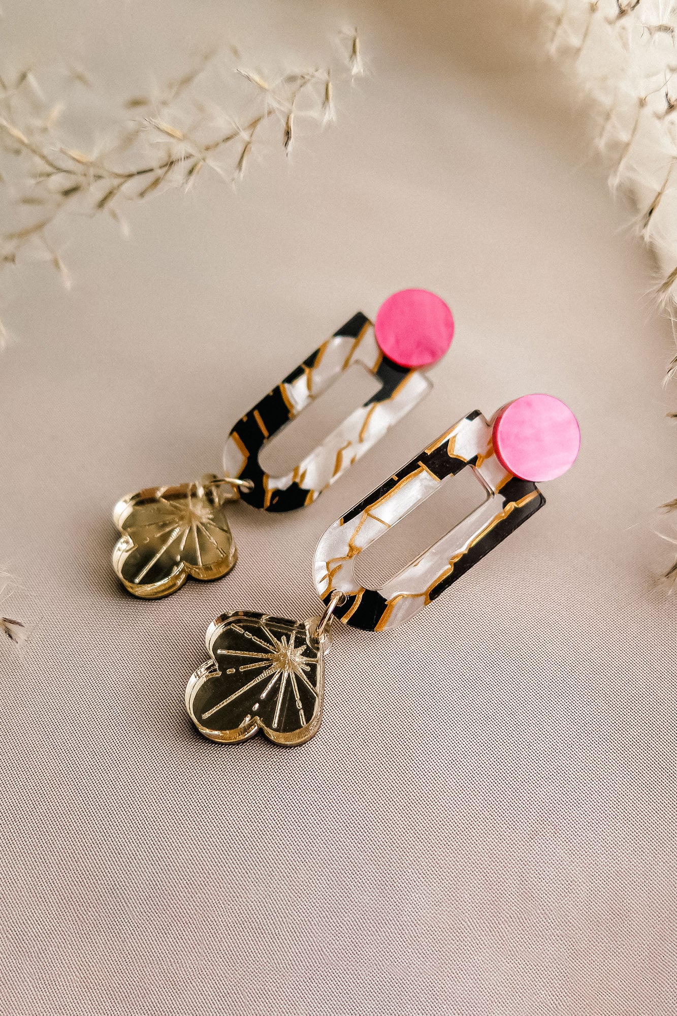 A pair of modern Sakura Earrings featuring pink circular studs, rectangular tortoiseshell links, and dangling gold heart-shaped charms with etched starburst designs, displayed on a light beige fabric background—an expression of artistry and elegance.