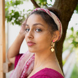 A woman with a light pink headscarf and gold Sakura Earrings stands outdoors near a tree, wearing a magenta top and looking at the camera with calm elegance. Green leaves and a white building are in the background.