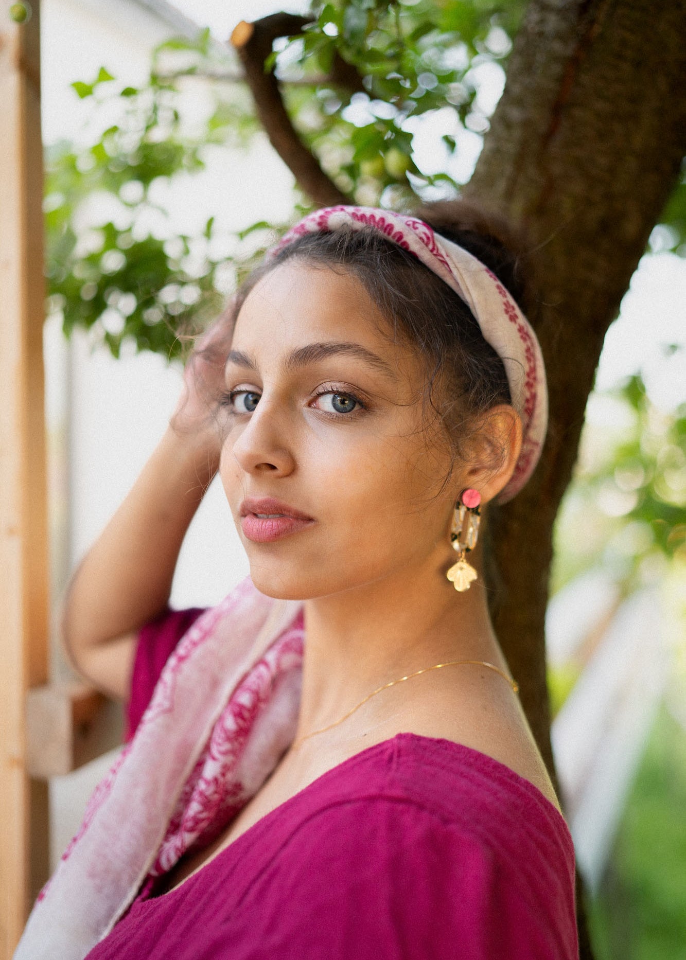 A woman with a light pink headscarf and gold Sakura Earrings stands outdoors near a tree, wearing a magenta top and looking at the camera with calm elegance. Green leaves and a white building are in the background.