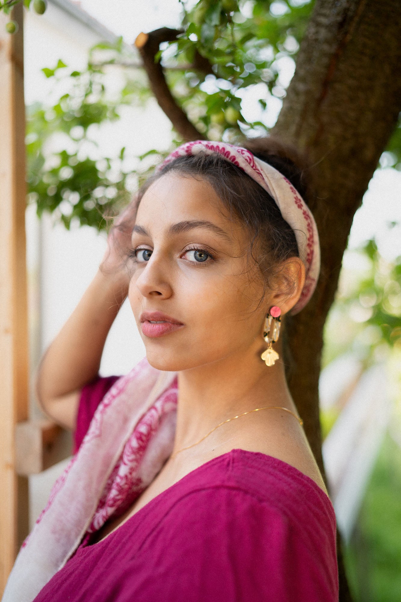 A woman with a light pink headscarf and gold Sakura Earrings stands outdoors near a tree, wearing a magenta top and looking at the camera with calm elegance. Green leaves and a white building are in the background.