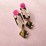 A pair of Sakura Earrings on a pink background, featuring pink circular studs, marbled rectangular links with gold accents, and clover-shaped gold charms at the bottom—a true blend of artistry and elegance.
