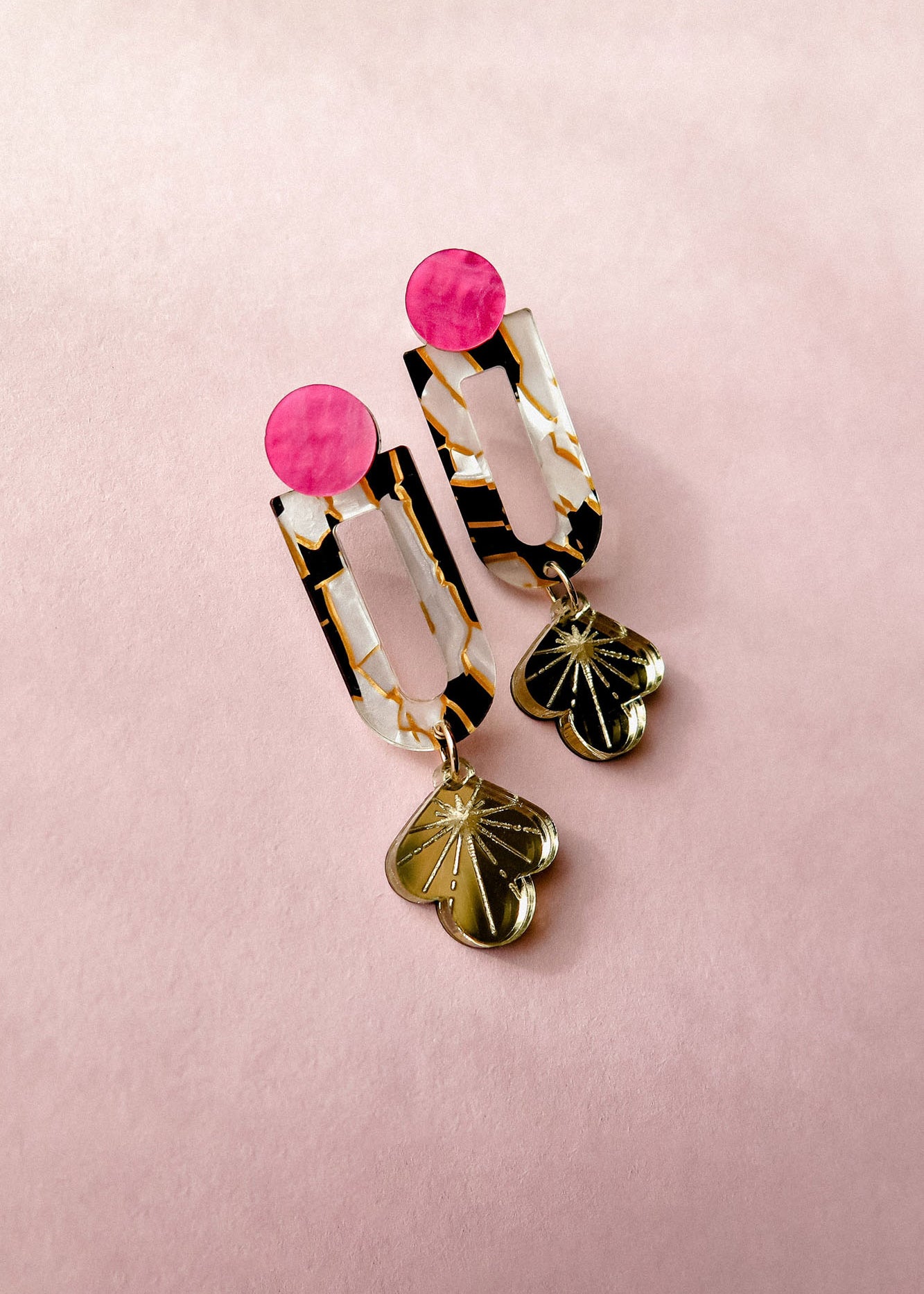 A pair of Sakura Earrings on a pink background, featuring pink circular studs, marbled rectangular links with gold accents, and clover-shaped gold charms at the bottom—a true blend of artistry and elegance.