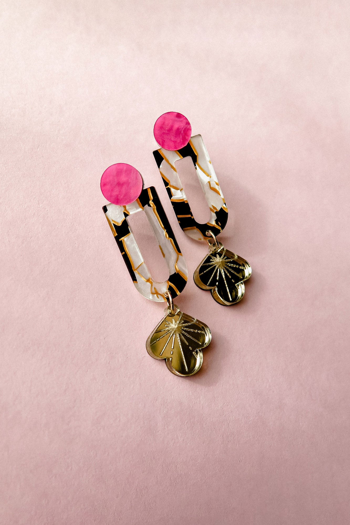 A pair of Sakura Earrings on a pink background, featuring pink circular studs, marbled rectangular links with gold accents, and clover-shaped gold charms at the bottom—a true blend of artistry and elegance.