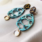 Statement earrings with geometric shapes: top hexagonal black and gold piece, middle turquoise open hexagon, and small gold oval dangle—these handmade accessories offer a touch of zero-waste beauty on a soft white fabric background.