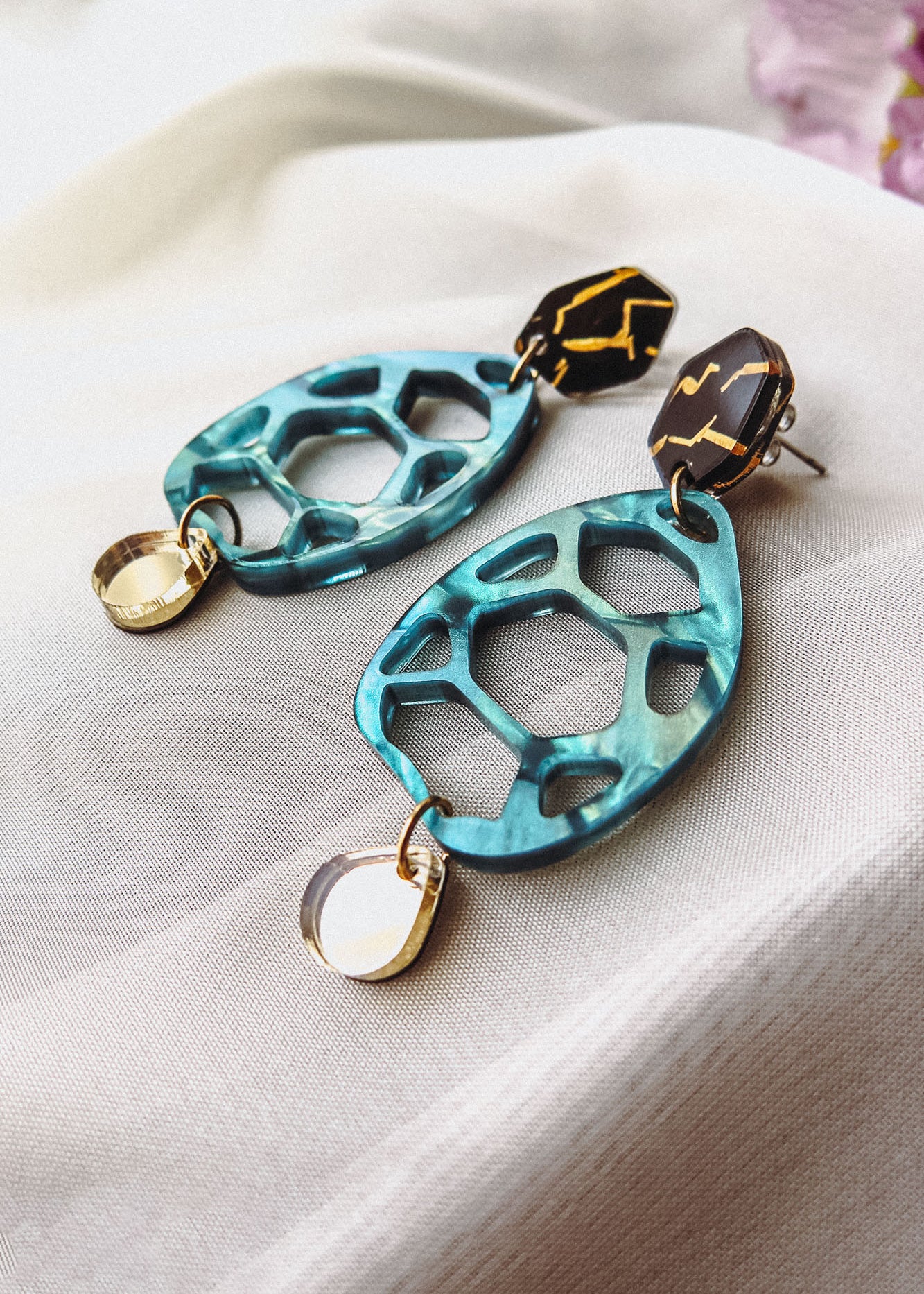 Statement earrings with geometric shapes: top hexagonal black and gold piece, middle turquoise open hexagon, and small gold oval dangle—these handmade accessories offer a touch of zero-waste beauty on a soft white fabric background.