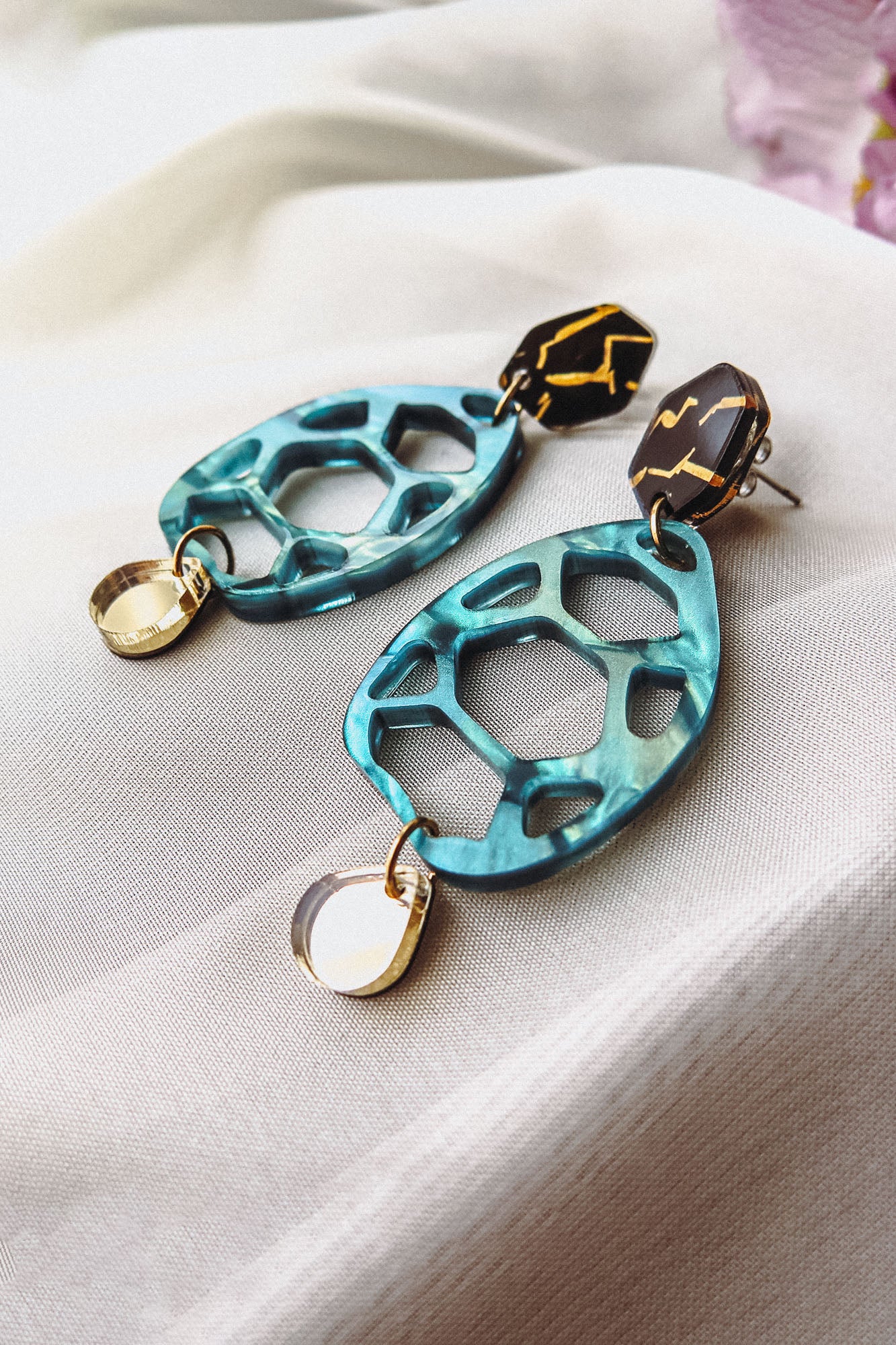 Statement earrings with geometric shapes: top hexagonal black and gold piece, middle turquoise open hexagon, and small gold oval dangle—these handmade accessories offer a touch of zero-waste beauty on a soft white fabric background.
