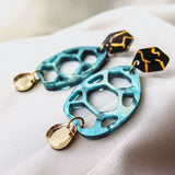 A pair of colorful drop earrings with geometric shapes—blue openwork teardrop, gold circular charm, and black hexagonal top—showcased as handmade accessories on a white fabric background.