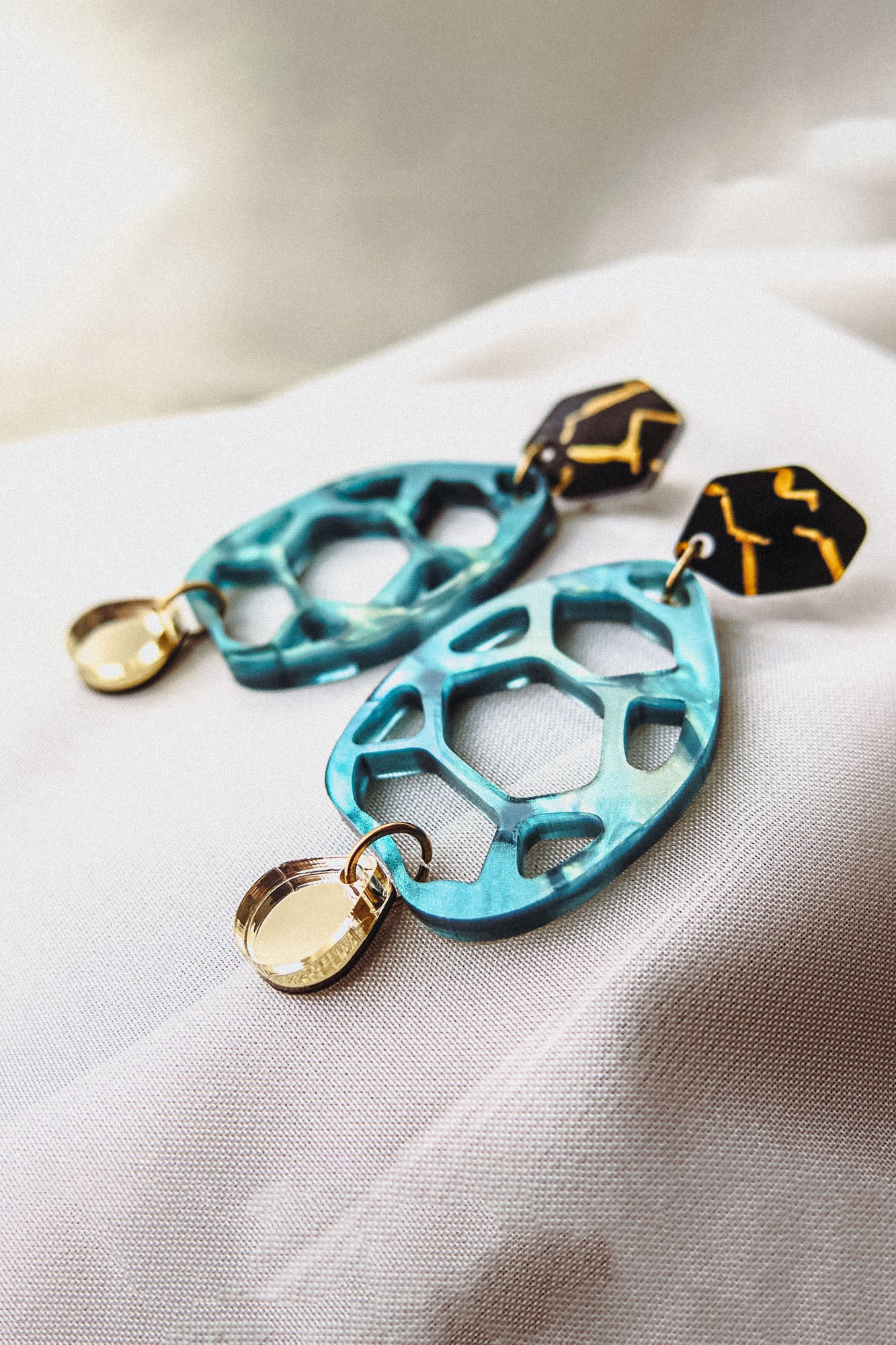 A pair of colorful drop earrings with geometric shapes—blue openwork teardrop, gold circular charm, and black hexagonal top—showcased as handmade accessories on a white fabric background.