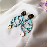 A pair of handmade sustainable earrings with black and gold geometric studs, turquoise oval-shaped openwork centers, and small gold circular charms, placed on white fabric with a pink flower in the corner.
