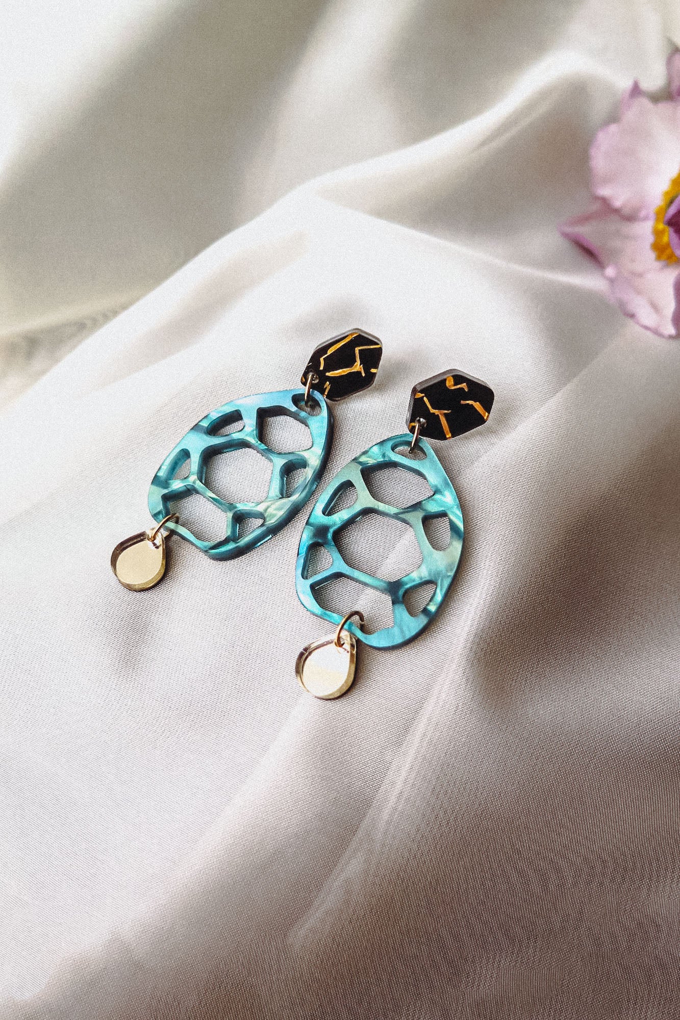 A pair of handmade sustainable earrings with black and gold geometric studs, turquoise oval-shaped openwork centers, and small gold circular charms, placed on white fabric with a pink flower in the corner.