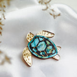A handcrafted sea turtle brooch with a gold and turquoise mosaic shell rests on smooth white fabric. Its fins are gold with brown spots, while delicate dried flowers blur the foreground, highlighting its artistic design.