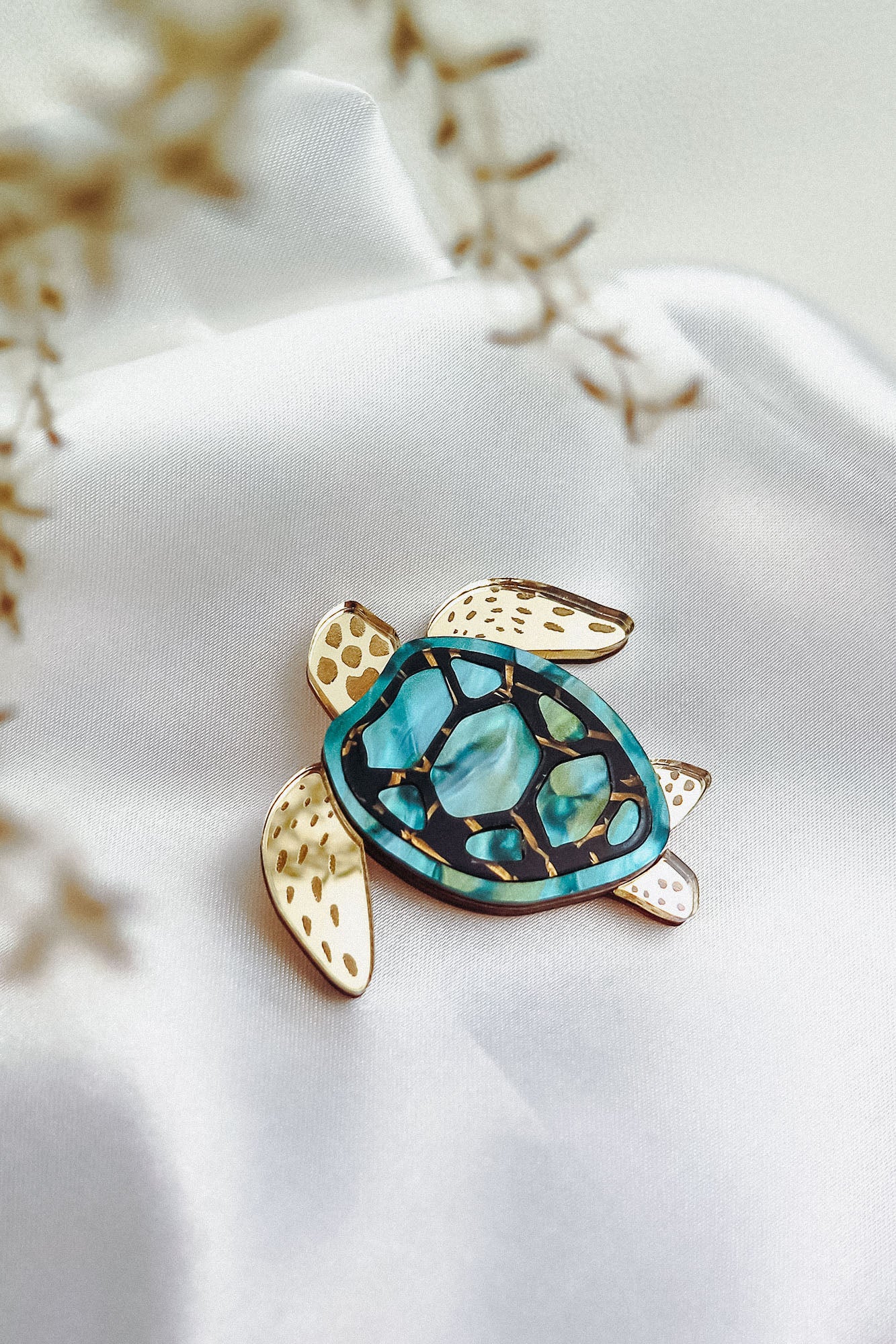 A handcrafted sea turtle brooch with a gold and turquoise mosaic shell rests on smooth white fabric. Its fins are gold with brown spots, while delicate dried flowers blur the foreground, highlighting its artistic design.