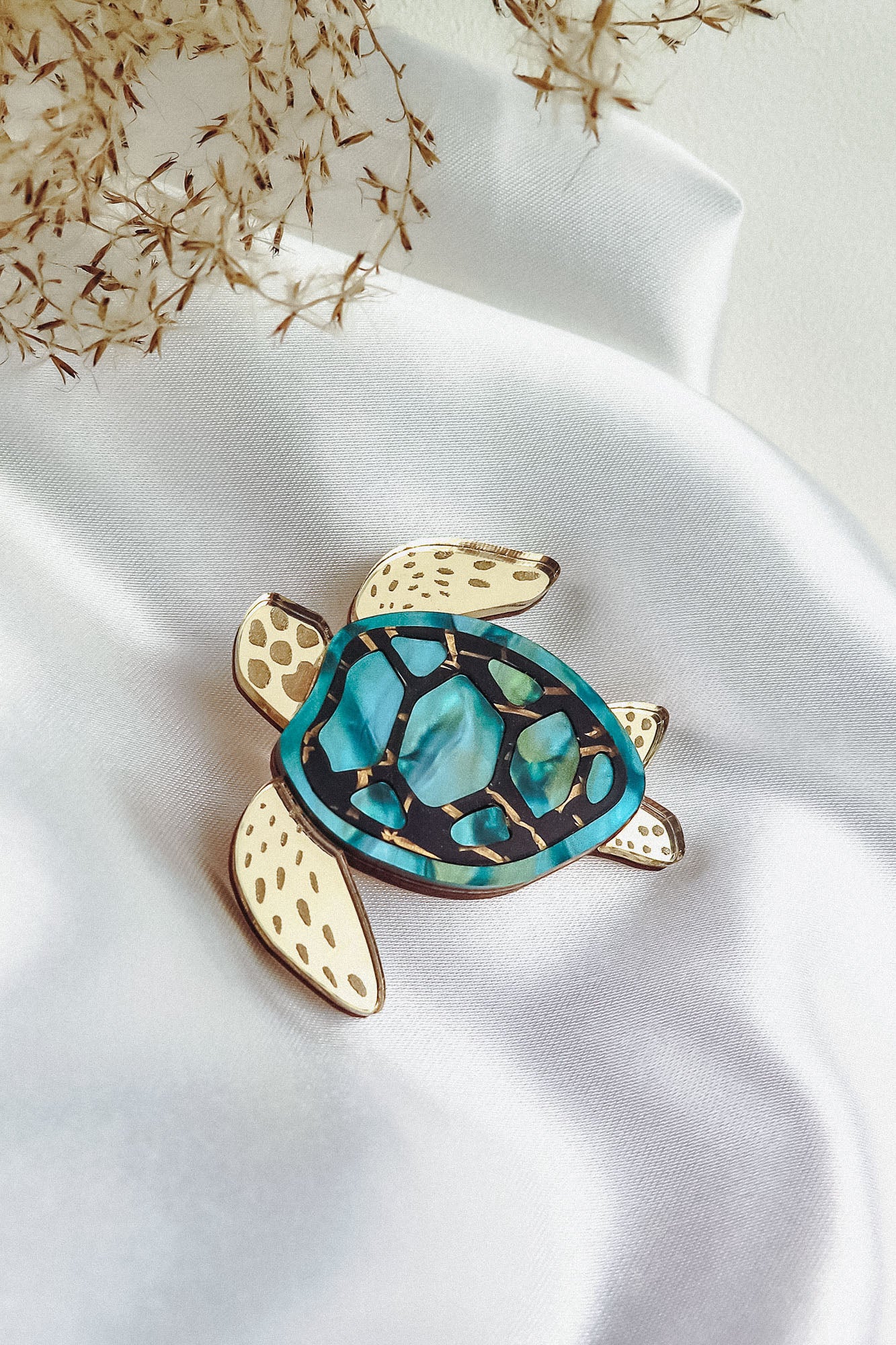 A handcrafted sea turtle brooch with a gold outline, cream-colored flippers, and a blue and green mosaic-patterned shell is displayed on white satin fabric with dried grasses nearby.