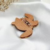 A handcrafted wooden bunny brooch with a metal pin clasp rests on white satin fabric, accented by delicate dried flowers in the top left corner, showcasing artistic design.