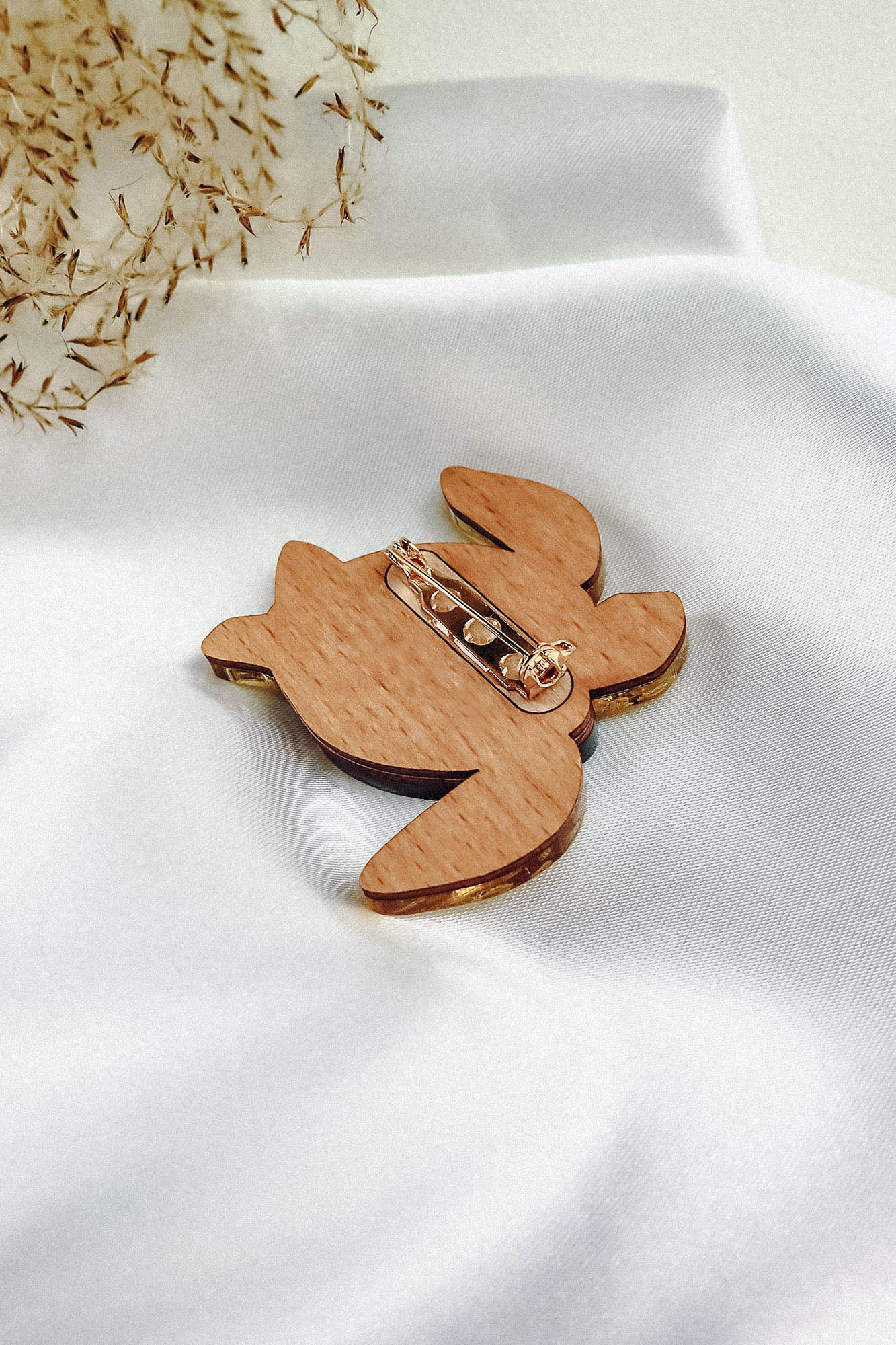 A handcrafted wooden bunny brooch with a metal pin clasp rests on white satin fabric, accented by delicate dried flowers in the top left corner, showcasing artistic design.